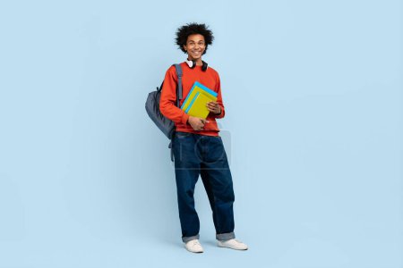 Casual dressed young african american man holding books with a backpack on a blue background