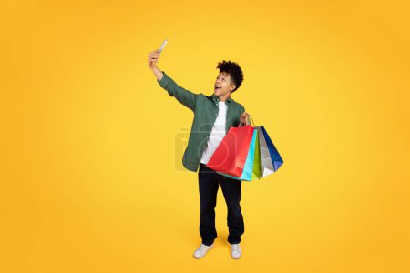 Téléchargez les photos : Riant homme afro-américain tenant des sacs à provisions et de prendre un selfie, exprimant la joie sur une toile de fond jaune - en image libre de droit