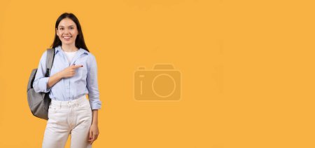 Photo for Attractive young woman with a backpack gestures and points to the side, looking away on an orange background - Royalty Free Image