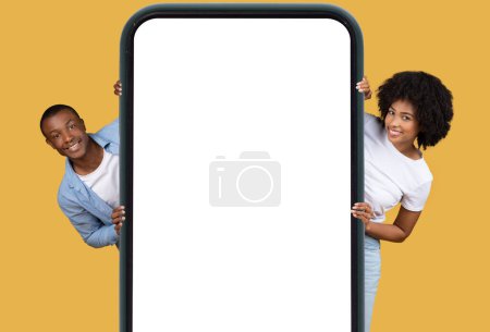 Foto de Pareja afroamericana sonriente que presenta una gran maqueta en blanco de la pantalla del teléfono inteligente sobre un fondo amarillo - Imagen libre de derechos