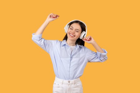 Cheerful woman with wireless headphones dancing and enjoying music, expressing happiness, yellow background