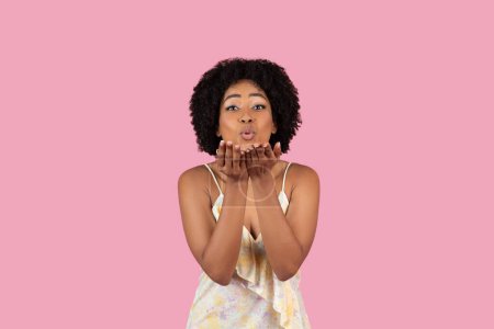 Junge Afroamerikanerin bläst einen fliegenden Kuss, ausdrucksstark und fröhlich auf rosa Studiohintergrund