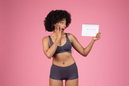 Gestresste junge Afroamerikanerin im Sport-BH schaut auf den Menstruationszyklus-Kalender und berührt ihr Gesicht vor rosa Hintergrund