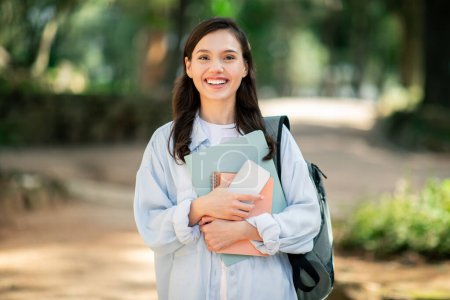 A joyful young woman student carrying a backpack and textbooks in a serene park setting, holding phone