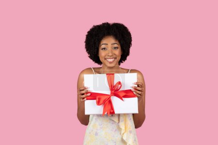 Téléchargez les photos : Une charmante jeune femme afro-américaine présentant fièrement une boîte cadeau blanche magnifiquement emballée avec un sourire - en image libre de droit