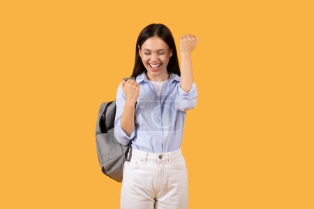 Photo for A joyful young woman with a backpack, clenching her fist in a celebratory gesture on a solid orange background - Royalty Free Image