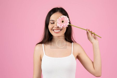 Foto de Una joven jovial pone juguetonamente una flor rosa gerbera cerca de su cara sobre un fondo rosa de estudio - Imagen libre de derechos