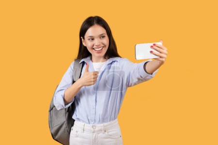 Eine fröhliche junge Frau mit Rucksack macht ein Selfie, lächelt fröhlich und zeigt eine Daumen-hoch-Geste auf gelbem Hintergrund.