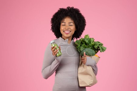 Foto de Contenido Mujer afroamericana sosteniendo una bolsa de verduras frescas y bebiendo jugo verde - Imagen libre de derechos