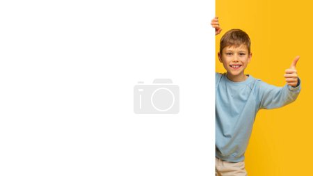 Joyeux jeune garçon avec un pouce vers le haut signe, moitié derrière un fond jaune avec de l'espace pour la maquette de texte
