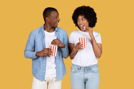 Photo for Engaged and cheerful African American couple enjoying popcorn and chatting on yellow, going to cinema - Royalty Free Image