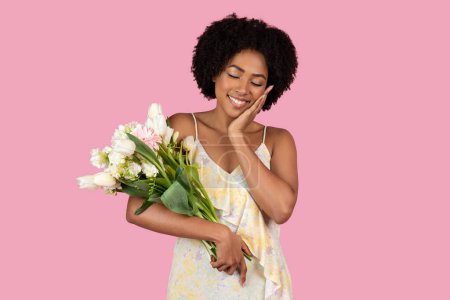 Téléchargez les photos : Une jeune afro-américaine en robe florale souriant tendrement, tenant un bouquet de fleurs fraîches sur fond rose, incarnant bonheur et gratitude - en image libre de droit