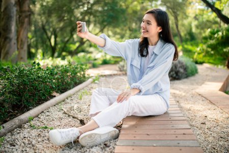 Mujer entusiasta capturar su propia foto con un teléfono inteligente en un entorno de parque representa la alegría de compartir en redes sociales
