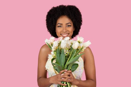 Foto de Hermosa mujer afroamericana sonríe sosteniendo un ramo de tulipanes blancos sobre un fondo rosa suave - Imagen libre de derechos