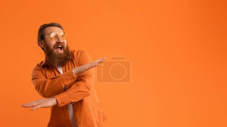 Photo for Red-haired man with beard dressed in bright shirt and sunglasses dances joyfully, crossing hands with excitement on orange studio background. Concept of positivity and cool party vibes. Panorama - Royalty Free Image