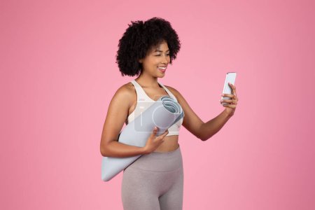 Téléchargez les photos : Femme afro-américaine souriante avec tapis de yoga engagé avec son smartphone - en image libre de droit