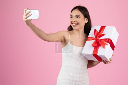 A cheerful woman in a white dress takes a selfie with a big white gift box with a red ribbon on a pink backdrop