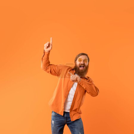 Photo for Energetic middle aged redhaired man with beard and sunglasses pointing upward with joy while dancing on orange studio background, radiates positive vibes, making funny moves. Copy space - Royalty Free Image