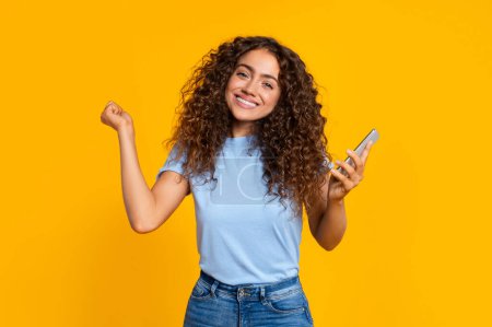 Fröhliche junge Frau hält Smartphone in der Hand und macht eine gewinnende Faustpumpe vor gelber, heller Kulisse