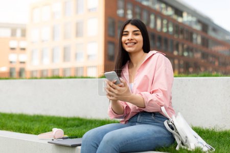 Ein Mädchen in einem lässigen rosafarbenen Hemd benutzt ihr Smartphone mit Laptop und Kopfhörer in der Nähe und lernt im Freien