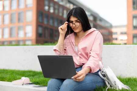 A young girl wearing glasses concentrates on her laptop screen while sitting outside in a grassy urban area