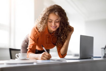 Foto de Mujer enfocada con el pelo rizado trabajando en su escritorio con un ordenador portátil y portátil, en un entorno de oficina brillante - Imagen libre de derechos