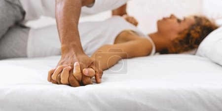 Téléchargez les photos : Moment intime de couple afro-américain tenant la main, mettant en valeur l'amour et le confort, sur un lit doux avec draps blancs - en image libre de droit