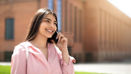Téléchargez les photos : Jeune étudiante engagée dans une conversation téléphonique, avec un ciel clair et le bâtiment de l'université en arrière-plan - en image libre de droit