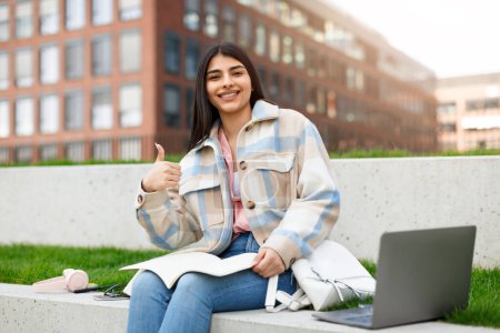 Foto de Una joven alegre se sienta en un césped del campus con su computadora portátil, libros de texto y auriculares mientras da un pulgar hacia arriba - Imagen libre de derechos