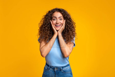 Photo for Cheerful curly-haired woman showing excitement and surprise, standing against a vibrant yellow backdrop - Royalty Free Image
