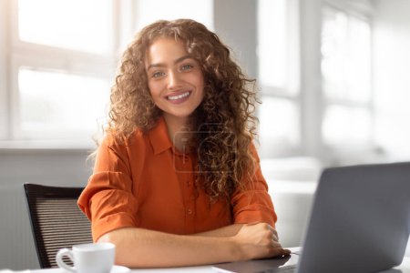 Téléchargez les photos : Jeune femme aux cheveux bouclés rayonne de bonheur alors qu'elle pose à son poste de travail lumineux avec ordinateur portable - en image libre de droit