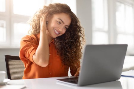 Eine strahlende junge Frau mit einem einnehmenden Lächeln berührt ihr lockiges Haar, ihren Laptop vor sich.