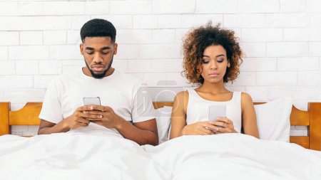 Foto de Estilo de vida de los Millennials. Joven afroamericano hombre y mujer ignorándose unos a otros, usando teléfonos en la cama, espacio libre - Imagen libre de derechos