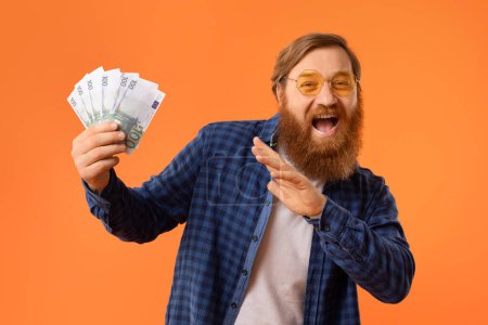 Photo for Positive mature redhaired man with beard holds euro banknotes in joyful celebration, shouting in victory, symbolizing a successful win in casino or lottery, against orange backdrop in studio - Royalty Free Image