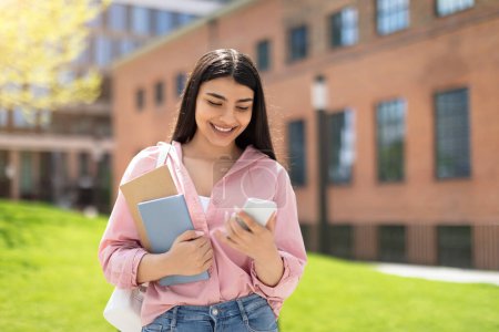 Foto de Una joven sosteniendo cuadernos sonríe mientras mira su teléfono inteligente con un edificio del campus en el fondo - Imagen libre de derechos