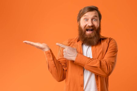 Téléchargez les photos : Homme excité avec des cheveux rouges et barbe pointant sur sa paume ouverte de la main, offre publicitaire ou produit, regardant l'appareil photo avec excitation sur fond orange, Studio Shot - en image libre de droit