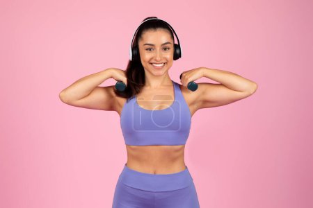 Téléchargez les photos : Une femme joyeuse portant des écouteurs et des vêtements d'entraînement fléchit ses muscles et sourit sur un fond rose - en image libre de droit