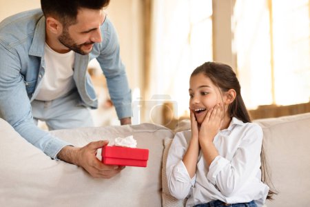A young man presents a red gift box to a delighted girl sitting on a sofa, capturing a moment of joy and surprise