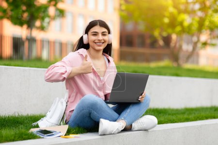 A content young girl sits with her laptop and headphones outdoors, giving a thumbs up to the camera