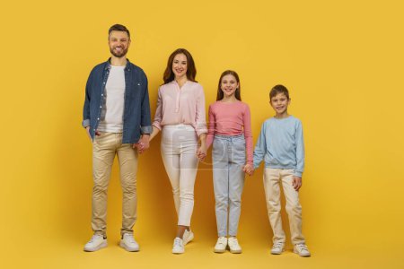 Affectionate family standing and smiling together on a yellow backdrop, father mother and kids holding hands