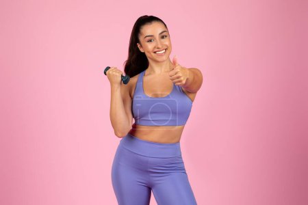 Confident woman in workout attire gives a thumbs up while holding dumbbells against a pink background