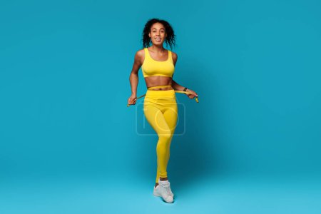 A happy, energetic African American woman in a yellow sports bra and leggings measuring waist against a vibrant blue background, representing health and vitality