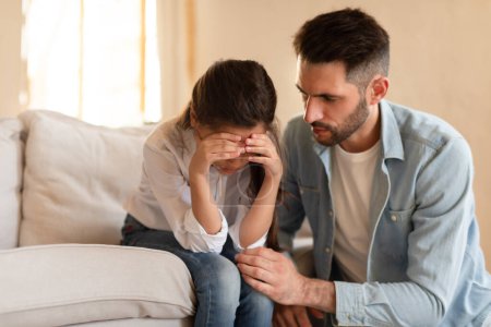 Loving father comforting crying daughter at home, man hugging his upset child sitting on couch, copy space