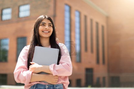 Téléchargez les photos : Étudiante joyeuse tenant un ordinateur portable fermé, regardant vers le haut avec réflexion à côté d'un bâtiment universitaire - en image libre de droit