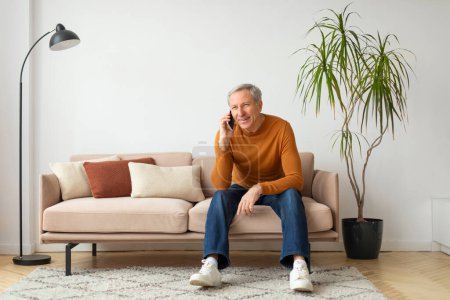 Photo for Cheerful mature man is seated on a beige couch in a well-lit room talking on a smartphone, copy space - Royalty Free Image