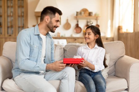Téléchargez les photos : Un père attentionné présente une boîte-cadeau rouge à sa jeune fille ravie assise sur un canapé à la maison - en image libre de droit