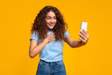 Junge Frau mit lockigem Haar sieht begeistert aus, während sie ein Selfie macht, isoliert vor gelbem Hintergrund