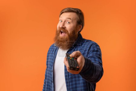 Photo for Cheerful redhaired bearded guy watching TV switching channels with remote control on orange backdrop. Portrait of happy middle aged man in casual enjoying movie on television, relaxing on weekend - Royalty Free Image