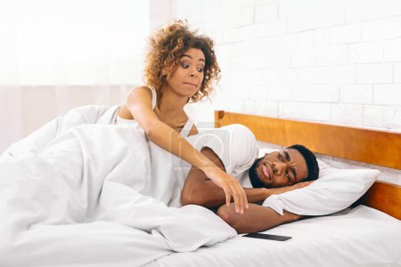 Hinterhältige schwarze Frau greift nach dem Telefon, während ihr Mann friedlich schläft, Unehrlichkeit und Betrug in Beziehungen
