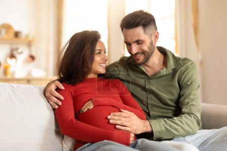 Photo for An expectant couple shares a tender moment on the sofa, with the focus on the pregnant womans belly - Royalty Free Image